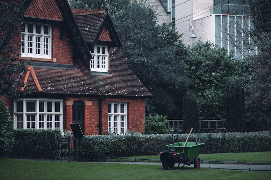 A traditional red brick house with a steep, tiled roof and multiple white-framed windows, set in a well-maintained garden. The garden features a neatly trimmed hedge bordering a lush green lawn, with