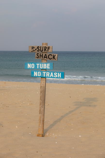 A wooden signpost on a sandy beach displaying three signs, with the top sign reading 'SURF SHACK' accompanied by a small palm tree icon, and the lower two signs stating 'NO TUBE' and 'NO TRASH' in bol